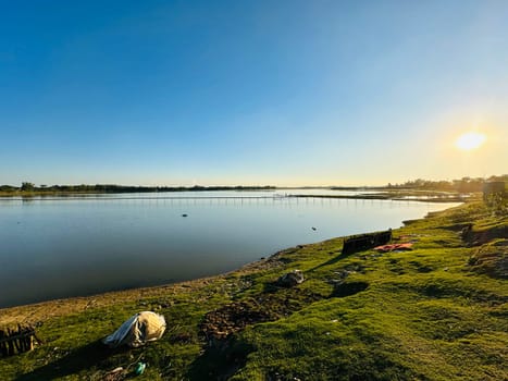 A tranquil lakeside scene featuring a vast expanse of water reflecting the golden glow of the setting sun. Lush green grass covers the bank, creating a picturesque backdrop. The clear blue sky extends across the horizon, adding to the serenity of the environment.