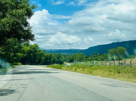 A scenic view of a winding asphalt road cutting through a lush green landscape, with dense trees lining the road on the left and a fence separating the road from a grassy field on the right. The road leads toward a distant mountain range under a clear blue sky dotted with white fluffy clouds.