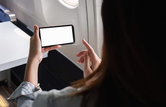 A young businesswoman using a smartphone with a blank screen while traveling by plane, highlighting modern technology in business travel.