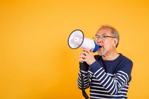 Portrait Asian old man wearing glasses screaming in megaphone studio shot isolated yellow background, happy elderly man holding a megaphone and shouts yelling announces news information