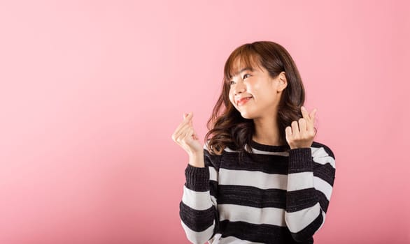 A confident woman stands with a smile, creating a mini heart sign with her finger on a pink background. Asian portrait of a beautiful young female sending love for Valentine's Day in a studio shot.