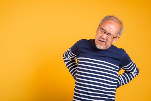 Portrait Asian old man with glasses holds hands on back he backache studio shot isolated yellow background, senior man pensioner suffering of backache, problems muscular pain