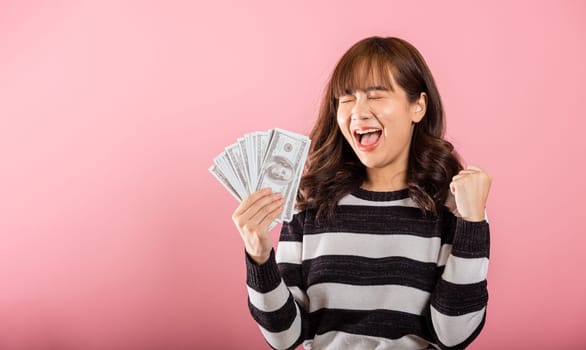 Asian happy portrait beautiful young woman standing smile her celebrating holding dollar money fan banknotes on hand and looking to camera on pink background with copy space for text