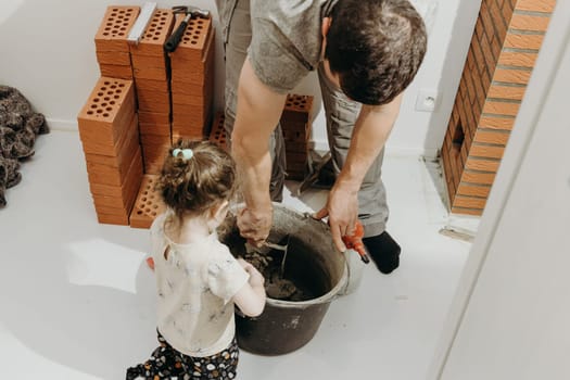 One young Caucasian man in uniform stands bent over and kneads cement mortar in a bucket with a spatula, his little daughter stands nearby and watches the process in the room where repairs and installation of a smoker for a fireplace made of bricks are underway, close-up side view from above. Concept of clean brickwork .