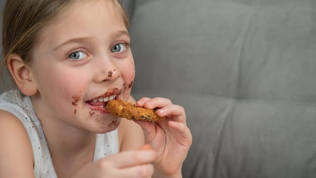 A cute little girl smeared in chocolate eats cookies while lying on the sofa