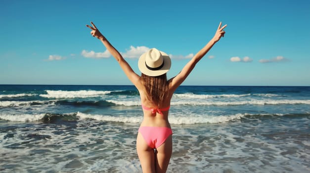 Summer vacation, rear view of beautiful happy slim woman in bikini swimsuit and tourist straw hat raising her hands up on the beach on sunny day on sea coast with waves