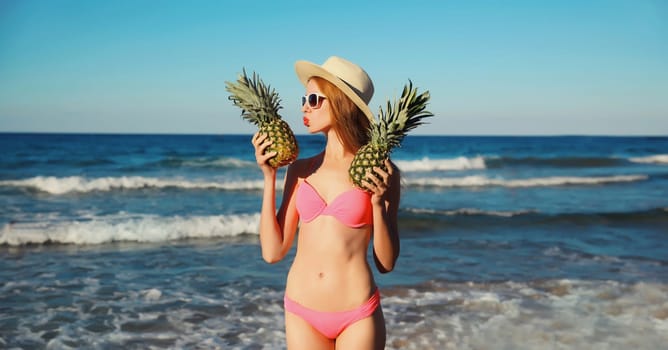 Summer vacation, beautiful happy young woman in pink bikini swimsuit, tourist straw hat, girl holding fruits kissing pineapple on the beach on sea coast with waves