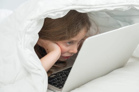 Portrait of a cute little girl using a laptop while lying under a blanket