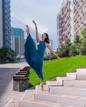 Beautiful Asian ballerina in a blue dress stands on the stairs in the splits outdoors. Urban landscape. Vertical photo