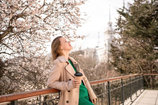 A woman is standing on a ledge with a cup of coffee in her hand. She is wearing sunglasses and a tan coat. The scene is set in a park with cherry blossoms in bloom. The woman is enjoying the view