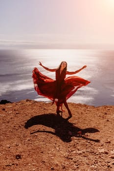 Woman red dress sea. Female dancer in a long red dress posing on a beach with rocks on sunny day. Girl on the nature on blue sky background