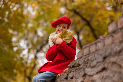 Smiling caucasian girl in a red coat and beret sits on a brick wall on a walk in autumn