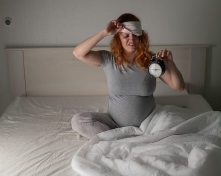 Caucasian pregnant woman sits in bed and holds an alarm clock. insomnia