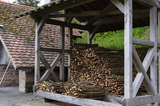 A neatly stacked pile of chopped firewood sits beneath a protective wooden structure. Sunlight filters through the slats in the roof, casting dappled shadows on the logs.