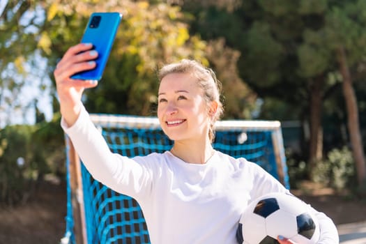 smiling young caucasian woman taking a selfie photo with mobile phone while holding a soccer ball in a urban field, concept of social media and football lifestyle