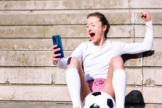 female football fan celebrates a victory when she is watching her mobile phone, concept of technology and urban soccer lifestyle, copy space for text
