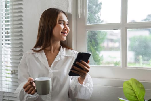 A beautiful woman drinks coffee in the morning while standing by a window in a bright, modern living room.