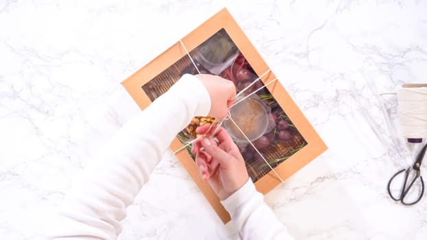 Flat lay. Hands are meticulously adding fresh red grapes to a bowl, complementing a beautifully arranged charcuterie box brimming with a variety of cheeses, olives, and cured meats, set against a sleek marble surface.