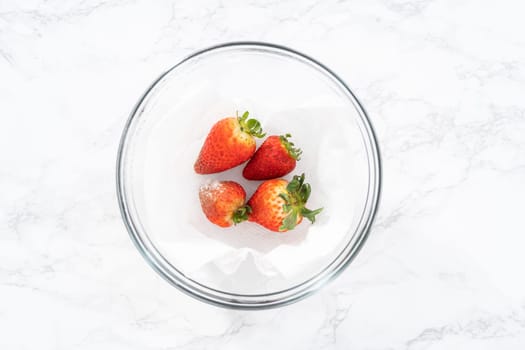 Bright red strawberries, interspersed with signs of mold, rest in a glass bowl lined with a paper towel on a white napkin, indicating improper storage techniques.