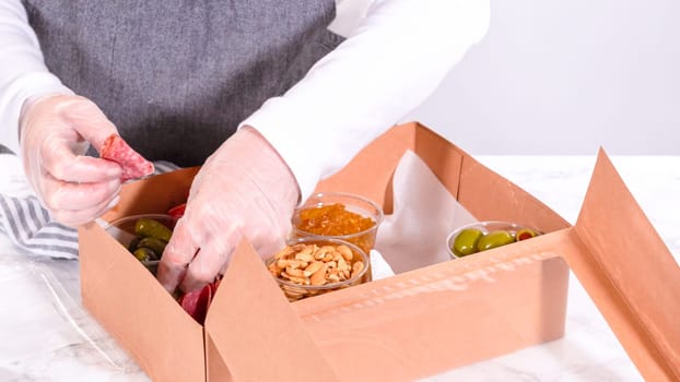 Hands are meticulously adding fresh red grapes to a bowl, complementing a beautifully arranged charcuterie box brimming with a variety of cheeses, olives, and cured meats, set against a sleek marble surface.