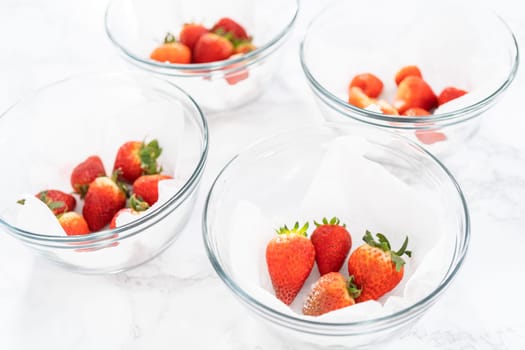 Bright red strawberries, interspersed with signs of mold, rest in a glass bowl lined with a paper towel on a white napkin, indicating improper storage techniques.