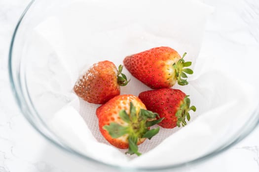 Bright red strawberries, interspersed with signs of mold, rest in a glass bowl lined with a paper towel on a white napkin, indicating improper storage techniques.