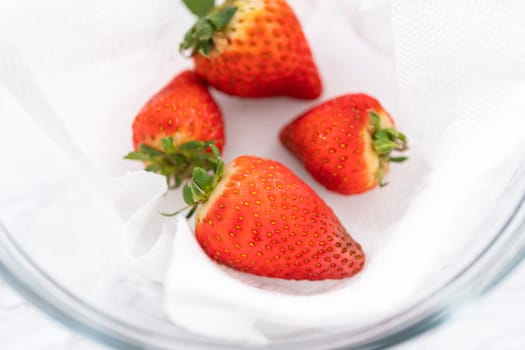 Bright red strawberries, interspersed with signs of mold, rest in a glass bowl lined with a paper towel on a white napkin, indicating improper storage techniques.