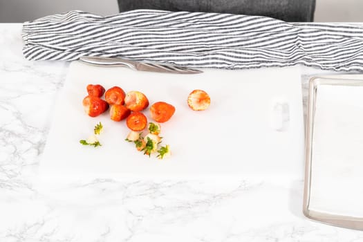 Bright, ripe strawberries are being carefully sliced on a white cutting board, a process that precedes drying or immediate consumption.