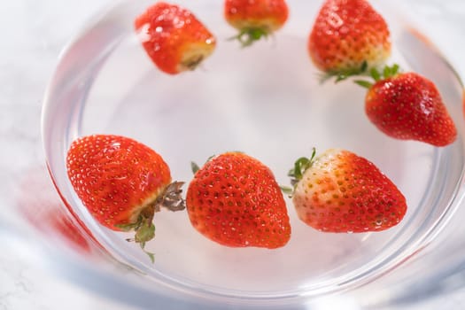 Ripe strawberries are submerged in water within a large glass mixing bowl, a step in washing the fruit to ensure cleanliness and longevity before storage or consumption.