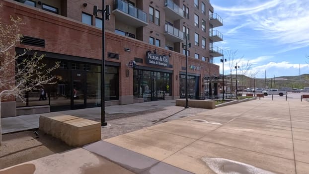 Castle Rock, Colorado, USA-June 12, 2024-Slow motion-A view of a modern brick building featuring retail shops and outdoor seating. The building architecture showcases large windows and balconies, adding to the urban ambiance. The spacious sidewalk and minimalistic decor reflect a clean and inviting environment in a bustling downtown area.