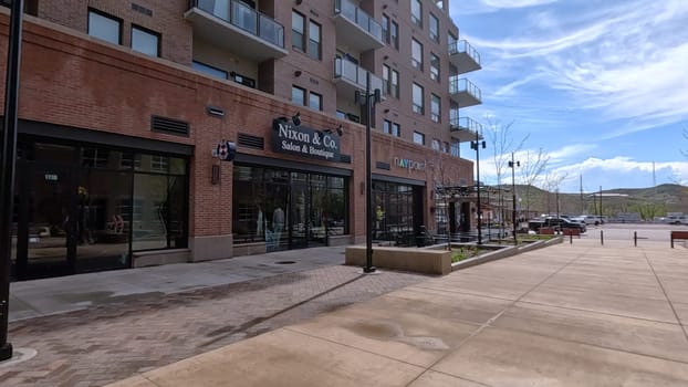 Castle Rock, Colorado, USA-June 12, 2024-Slow motion-A view of a modern brick building featuring retail shops and outdoor seating. The building architecture showcases large windows and balconies, adding to the urban ambiance. The spacious sidewalk and minimalistic decor reflect a clean and inviting environment in a bustling downtown area.