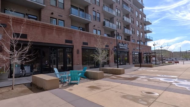 Castle Rock, Colorado, USA-June 12, 2024-Slow motion-A view of a modern brick building featuring retail shops and outdoor seating. The building architecture showcases large windows and balconies, adding to the urban ambiance. The spacious sidewalk and minimalistic decor reflect a clean and inviting environment in a bustling downtown area.