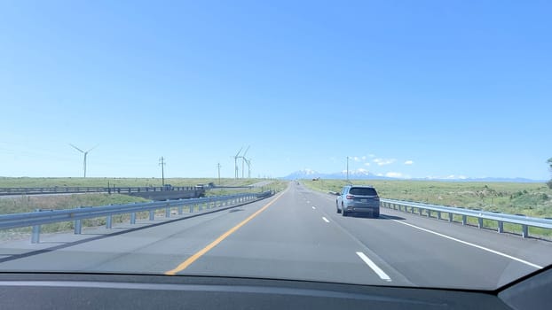 Pueblo, New Mexico, USA-June 10, 2024-Slow motion-A scenic highway drive featuring wind turbines dotting the landscape and a clear view of distant mountains. The image captures the open road on a sunny day, with a few vehicles traveling along the well-maintained highway. The bright blue sky and expansive green fields create a sense of calm and freedom.