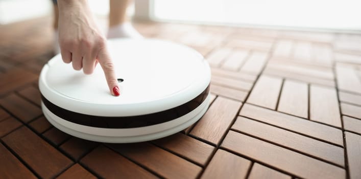 Female finger presses power button on white robotic vacuum cleaner. Home cleaning appliances concept