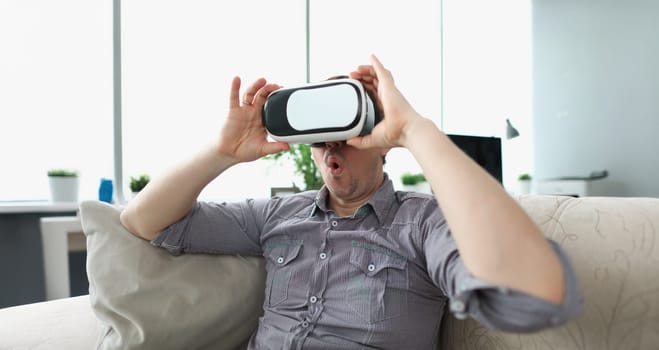 Surprised man wearing virtual reality glasses sitting on couch at home. Home entertainment concept