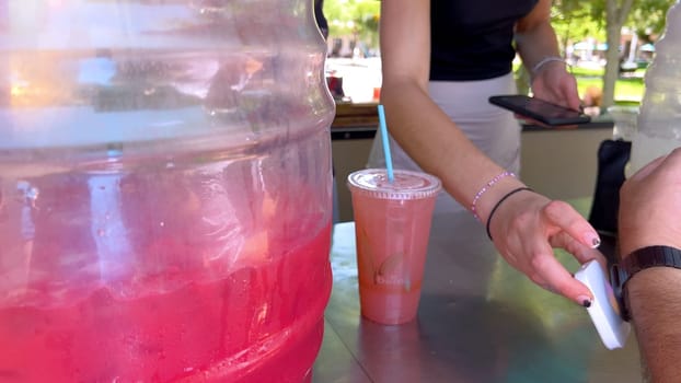 Santa Fe, New Mexico, USA-June 11, 2024-Slow motion-A close-up of a customer using an Apple Watch to pay for a refreshing drink at an outdoor juice stand. The image captures the transaction in progress, with a clear view of the large container of pink juice and the freshly prepared drink in a plastic cup with a straw. The scene is set in a sunny, vibrant outdoor market.