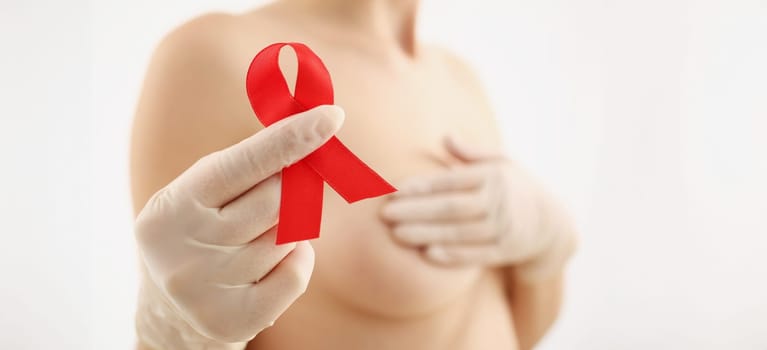 Young woman holding red ribbon in her hand and covering her breasts closeup. Prevention of breast cancer concept