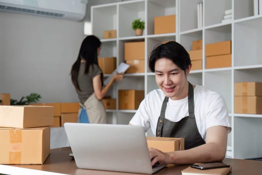 A couple managing their online business, taking orders, and packing products in a home office filled with cardboard boxes and a laptop.