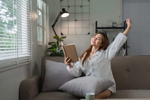 A young woman sits comfortably on a sofa, reading a book and stretching in the morning light, creating a serene and peaceful atmosphere.