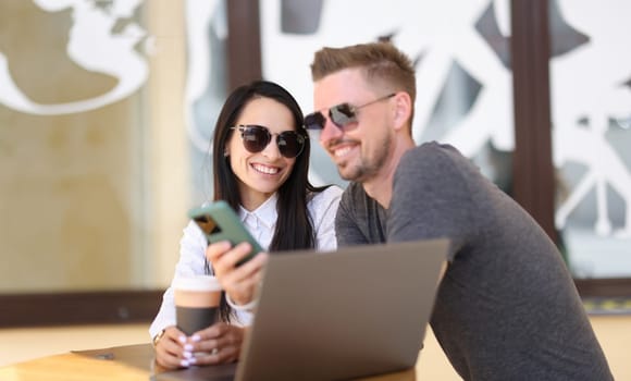Smiling man and woman in sunglasses communicate in coffee near laptop. Business meeting and communication concept