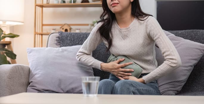 A young woman sits on a couch, holding a heating pad to her abdomen, experiencing menstrual cramps in a cozy living room setting.