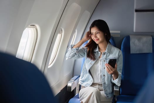 A young businesswoman travels by plane, relaxing in her seat, holding a smartphone, and looking out the window. Perfect for themes of business travel and modern professional life.
