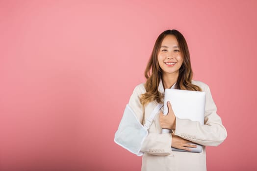 Asian business woman confident smiling broken arm after accident and wear arm splint for treatment but still have to work she hold laptop computer studio isolated on pink background,copy space