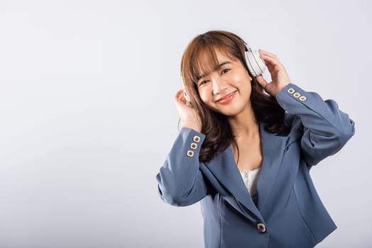 Happy Asian young female, wearing Bluetooth headphones, smiles while listening to music on her phone. Studio shot, isolated on white. She's fully engaged with modern technology and modern lifestyle.