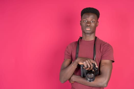 A man in a red shirt is holding a camera. He is standing in front of a pink wall