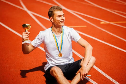 A young athlete sitting on field, holding trophy and gold medal, smiling in victory after winning a race