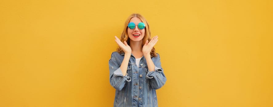 Portrait of stylish happy surprised young woman, cheerful girl in glasses, denim jacket posing on bright orange wall background