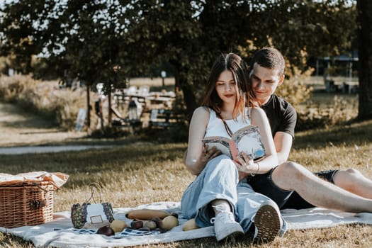 One beautiful Caucasian happy brunette girl sits on a blanket with a basket and fruits in the park on a picnic and reads a book while a guy hugs her waist on a summer day against the background of blurred trees, close-up side view.