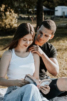 One beautiful Caucasian happy brunette girl sits on a blanket with a basket and fruits in the park on a picnic and reads a book, and the guy holds her by the shoulders and looks tenderly at her on a summer day against the backdrop of a blurred lawn, side view close-up.