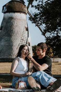 One beautiful Caucasian happy brunette couple sits full-length on a blanket with a basket and fruits in the park on a picnic and the guy treats the girl with melted juicy grapes on a summer day against the backdrop of an old windmill, close-up side view.